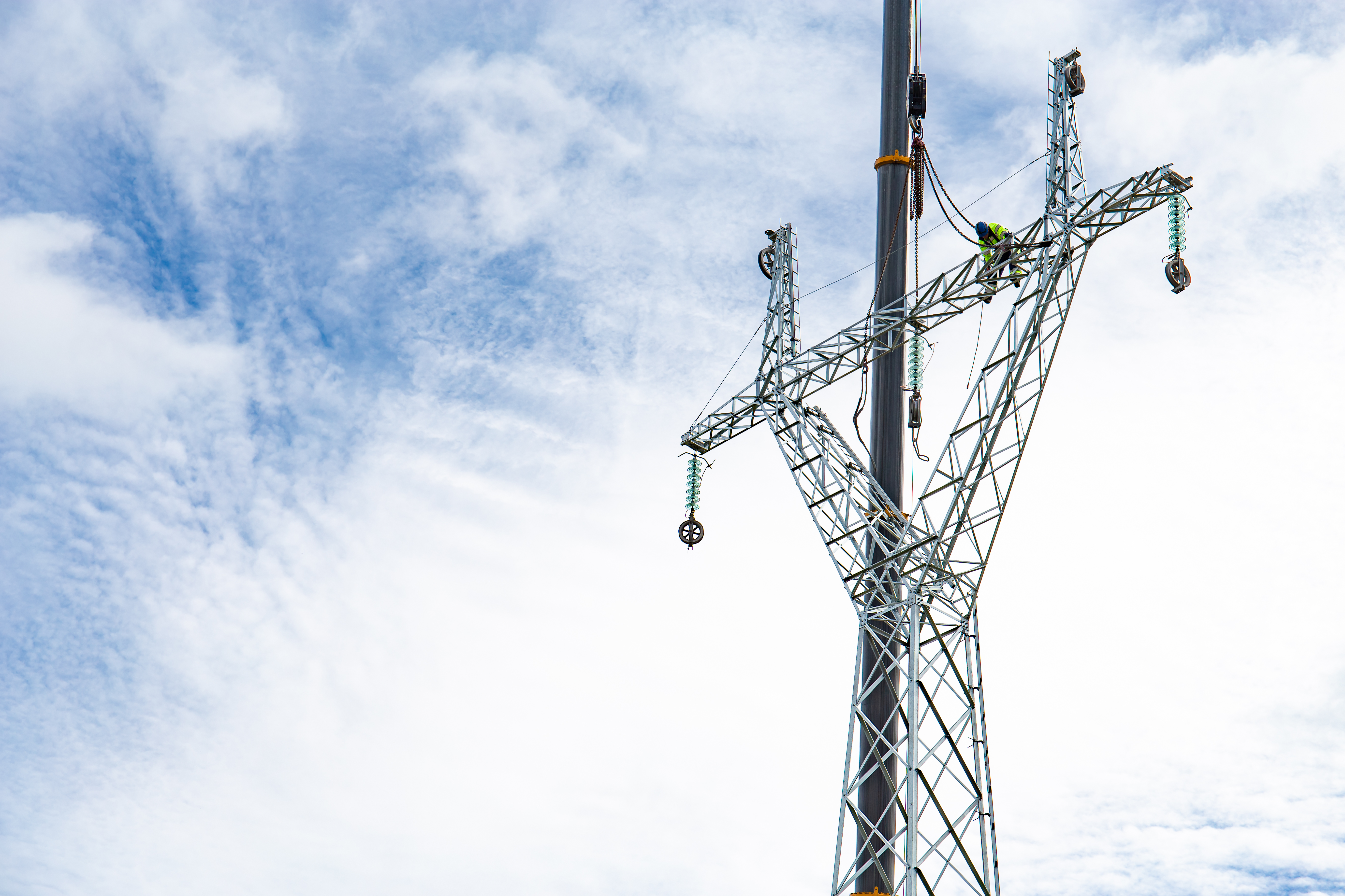 An image showing a tall metal utility structure with a worker positioned high on the framework. The structure resembles part of an electrical transmission tower, with insulators and cables attached. The sky in the background is bright and partly cloudy.