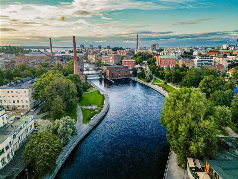 A view of Tampere with a river flowing through the center, buildings, trees, and bushes on both sides, under a partly cloudy sky.