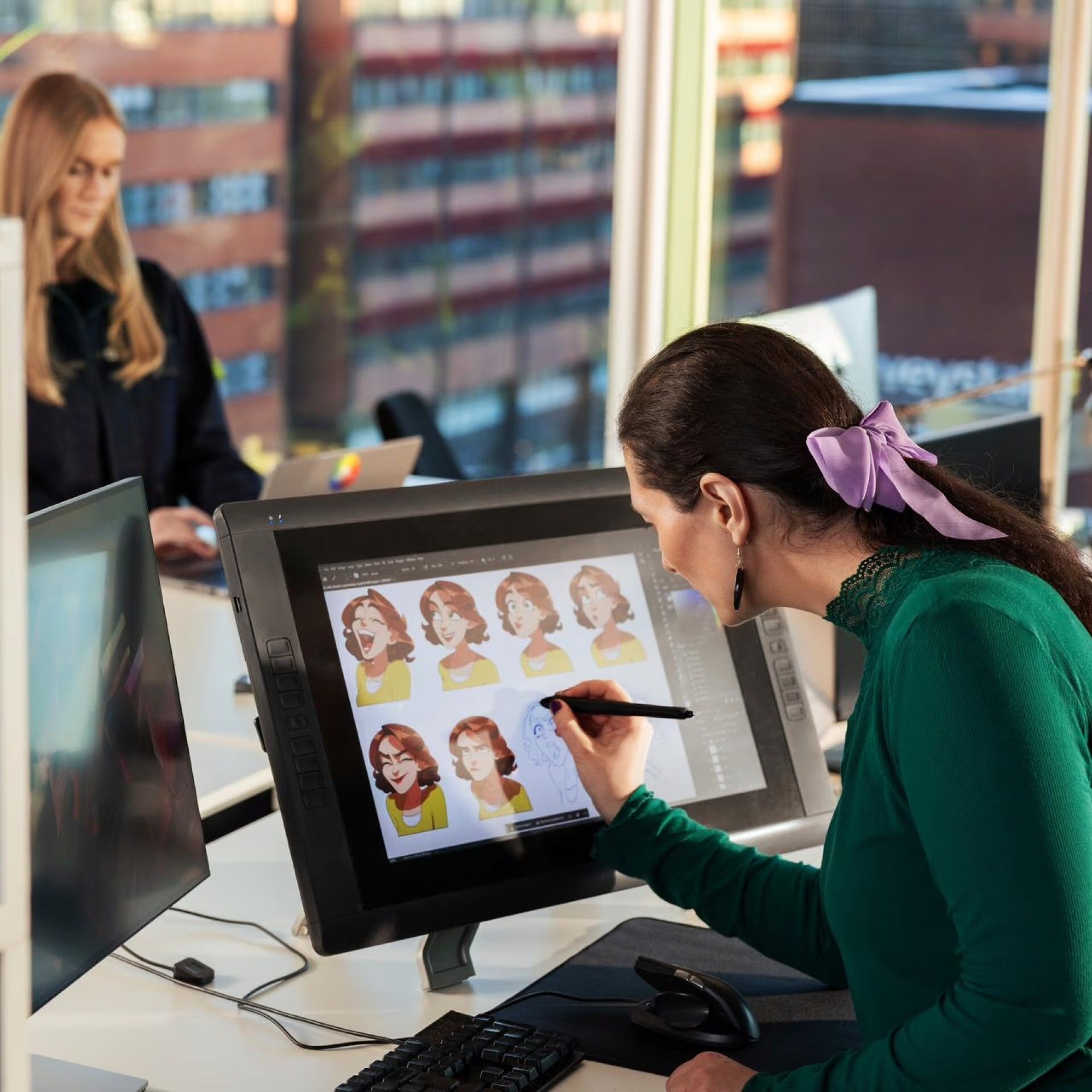 A digital artist drawing character expression sheets on a drawing tablet with a stylus in a modern office environment, with a colleague working in the background.