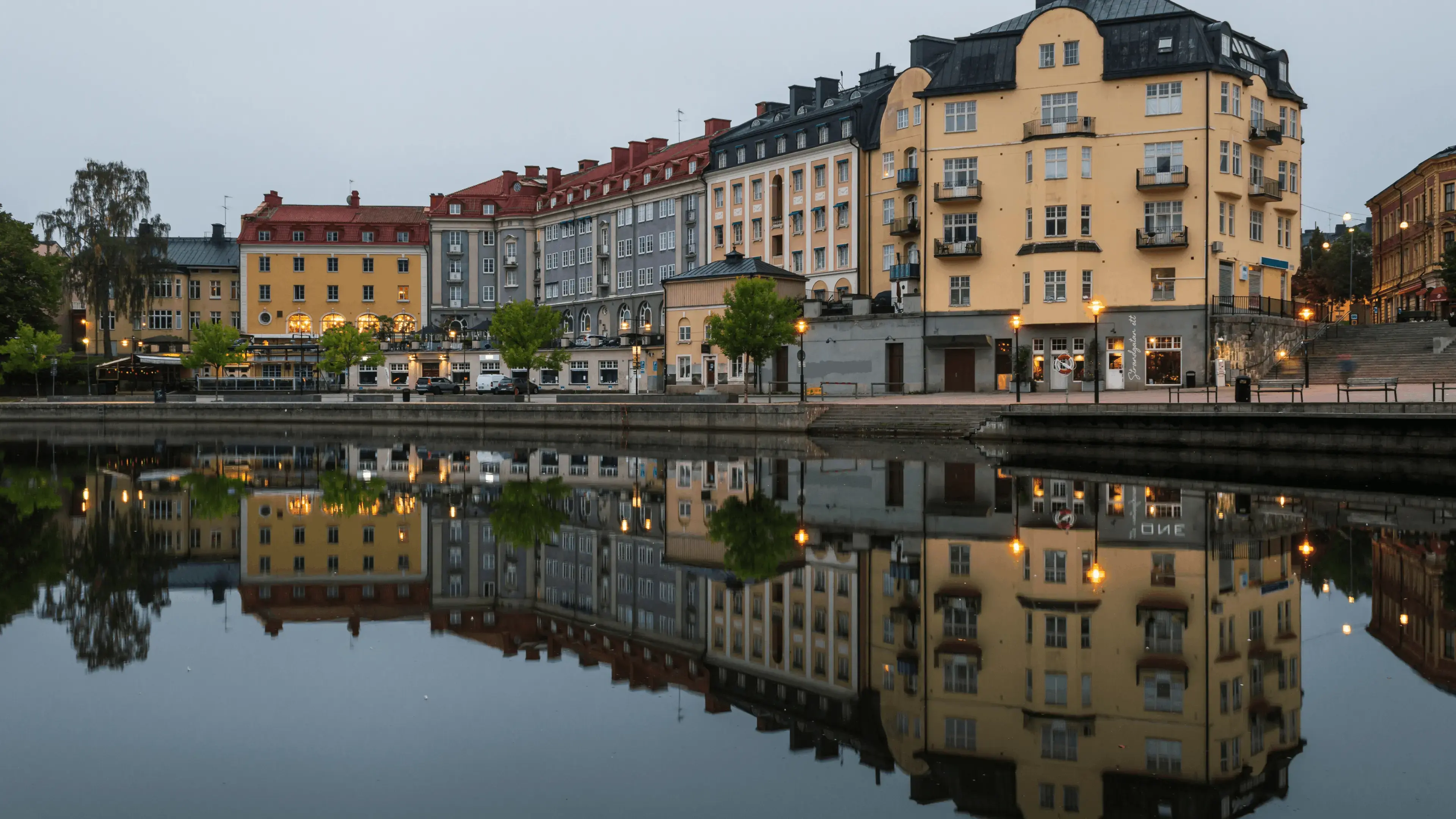 Waterfront view with multi‑story buildings reflected in calm water. Residential and commercial buildings line the quay, with illuminated streetlights and storefronts visible along the shoreline.