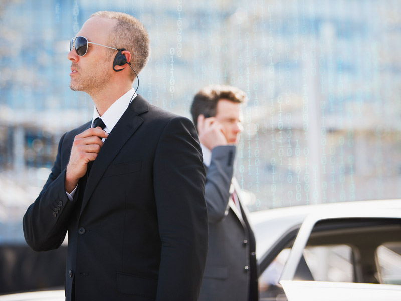 Two people stand outdoors near a vehicle, wearing formal clothing and earpieces. One person adjusts clothing at the neckline while the other speaks into a handheld device. The background shows an urban setting with glass structures.