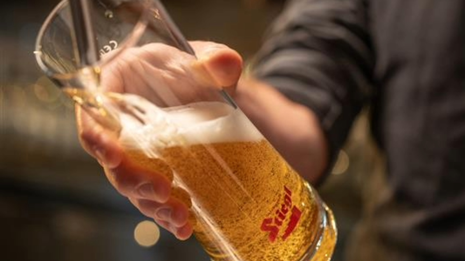 A hand holding a branded Stiegl beer glass filled with golden beer and a frothy white head, with a warm, blurred background suggesting a bar or brewery setting.