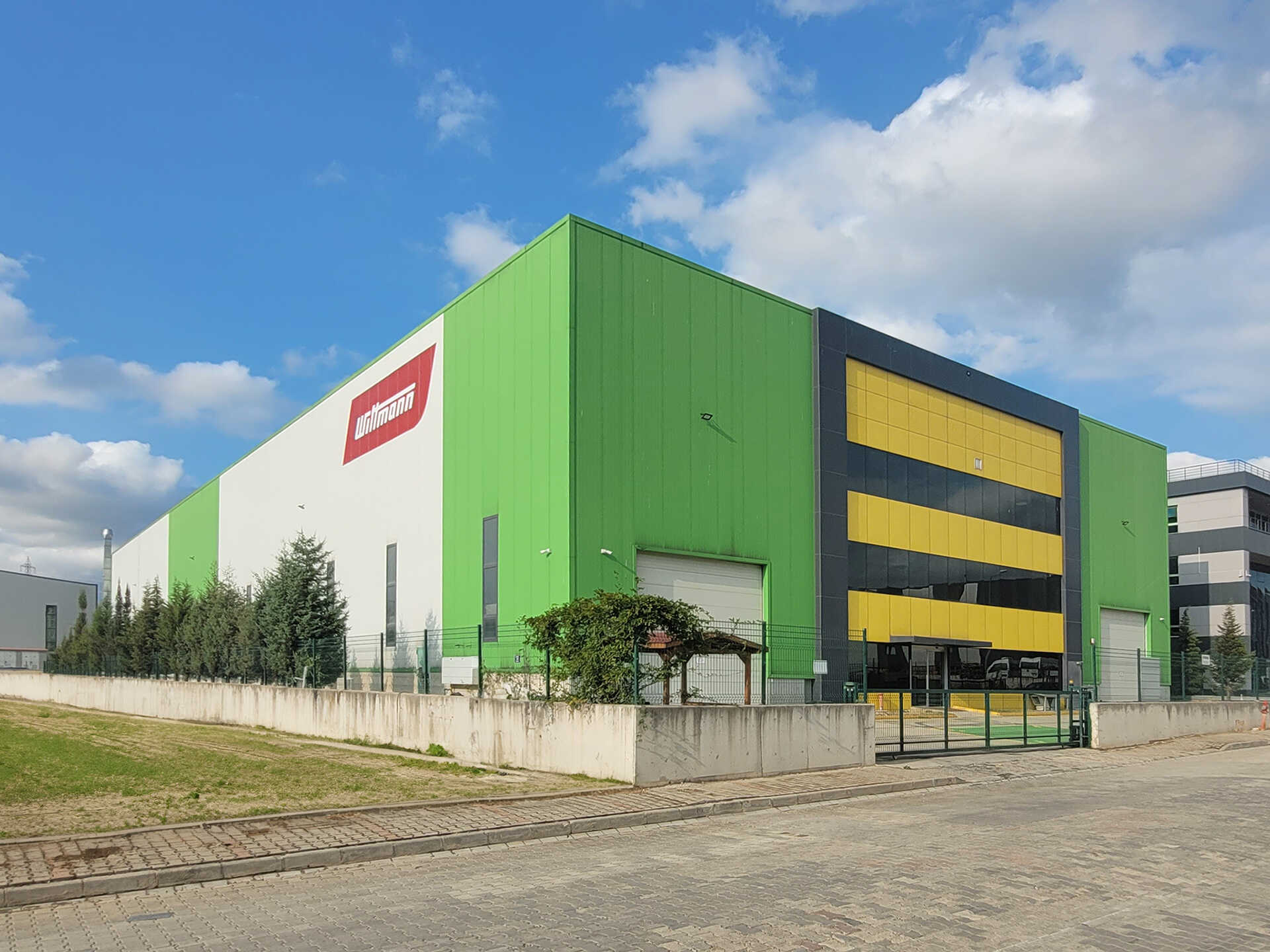 Exterior view of a large industrial building with green façades and yellow‑and‑black vertical window panels. The building stands in an industrial area with a paved road, fencing, and a partly cloudy sky.